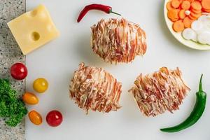 Potato filled with bacon ready to be baked in oven. Cheese, carrot, peppers some onion, tomato and herbs on table. photo