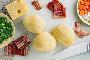 Sliced potato and bacon with knife,potato ready to be filled with bacon. Cheese, carrot some onion and herbs on table. photo