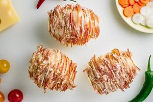 Potato filled with bacon ready to be baked in oven. Cheese, carrot, peppers some onion, tomato and herbs on table. photo