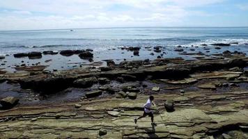 foto de rastreamento de vista lateral de um jovem correndo na costa de uma praia rochosa do oceano.