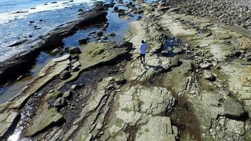 Tracking shot of a young man running on a rocky ocean beach shoreline.