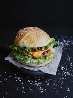 apetitosa hamburguesa casera fresca, con chuleta de pollo, lechuga, tomate, queso y salsa. sobre una tabla de madera sobre un fondo negro foto