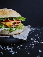 apetitosa hamburguesa casera fresca, con chuleta de pollo, lechuga, tomate, queso y salsa. sobre una tabla de madera sobre un fondo negro foto