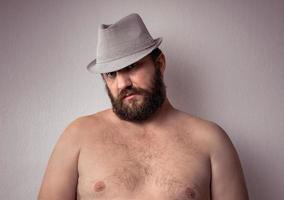 apuesto hombre barbudo semidesnudo con sombrero gris de pie contra la pared de fondo gris. foto