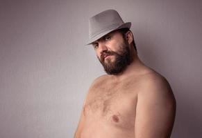 apuesto hombre barbudo semidesnudo con sombrero gris de pie contra la pared de fondo gris. foto