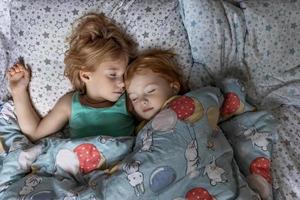 Two little sibling girls sisters sleeping in an embrace in bed under one blanket photo