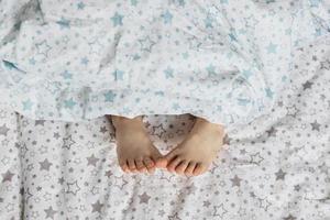 primer plano de los pies de la niña pequeña en la cama debajo de la manta. Tonos celestes y beige. foto