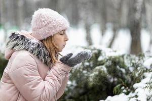 Mujer feliz en un día nevado de invierno en el parque, vestida con ropa de abrigo, sopla la nieve de sus guantes
