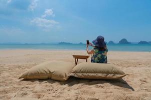Mujer joven sentada en un columpio cerca de la playa y usando un teléfono inteligente para tomar fotos durante las vacaciones de verano