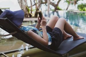 Mujer joven con sombrero y ropa casual sentada en el banco cerca de la piscina y usando un teléfono inteligente para tomar fotos durante las vacaciones de verano