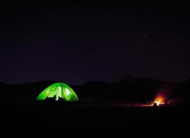 carpa verde iluminada de pie junto a la fogata en la naturaleza por la noche con fondo de noche estrellada. foto