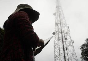 Female engineer using tablet computer checking the frequency with telecommunication towers with TV antennas and satellite dish In the foggy morning. photo