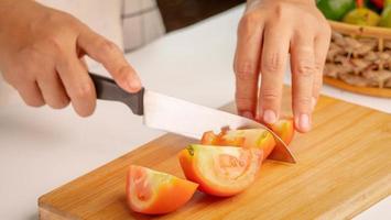 mujer cortando tomate fresco en trozos con un cuchillo sobre una tabla de cortar de madera.
