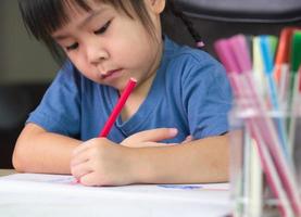 A cute little girl is drawing a rainbow on paper with colored pencils at the table at home. photo