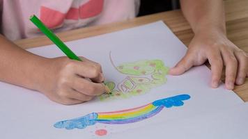 Cerrar la mano del niño dibujando un arco iris y un árbol con lápices de colores en la mesa en casa. foto