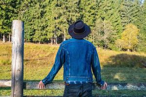 vaquero en una chaqueta de mezclilla y un sombrero cerca de la valla en las montañas foto