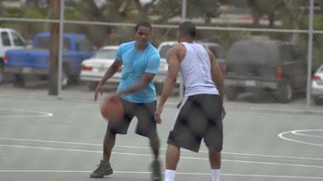 twee jonge mannen die samen een-op-een basketbal spelen op een buitenbaan omringd door een gaashekwerk.