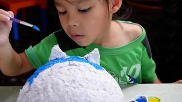 A little girl paints with a brush on the figure of a white pig-shaped papier-mache at home. Make your own piggy bank. video