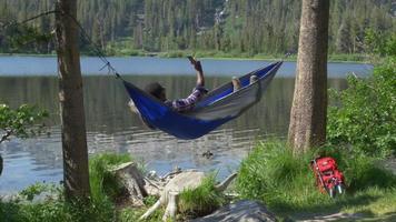 A man taking a selfie with his mobile device while resting in a hammock near a mountain lake. video