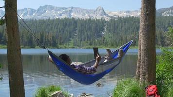 un hombre descansando en una hamaca y usando un dispositivo móvil tableta cerca de un lago de montaña.