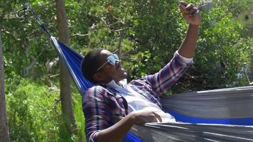 un hombre que se toma una selfie con su dispositivo móvil mientras descansa en una hamaca cerca de un lago de montaña. video
