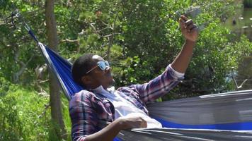 un hombre que se toma una selfie con su dispositivo móvil mientras descansa en una hamaca cerca de un lago de montaña. video