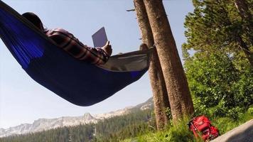 un uomo che riposa su un'amaca e utilizza un dispositivo mobile tablet vicino a un lago di montagna. video