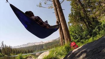 un uomo che riposa su un'amaca e utilizza un dispositivo mobile tablet vicino a un lago di montagna. video
