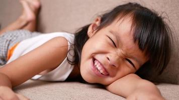 Cute little girl lying on sofa in the living room, looking at camera and smiling while playing at home. video