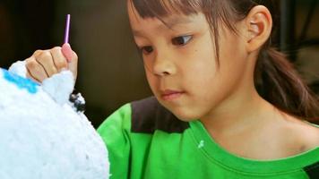 A little girl paints with a brush on the figure of a white pig-shaped papier-mache at home. Make your own piggy bank. video