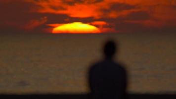 Silhouette eines Mannes, der den Sonnenuntergang am Strand beobachtet. video