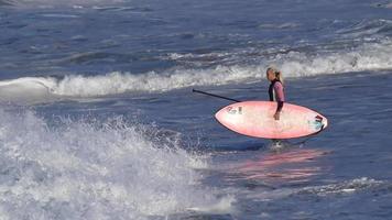 uma mulher monta uma prancha sup stand up enquanto surfa em uma prancha rosa.