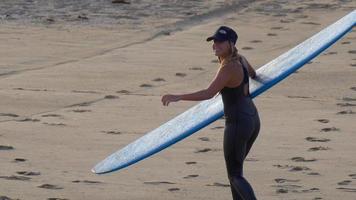 une jeune femme en combinaison de plongée marchant avec sa planche de surf longboard.
