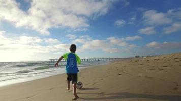 een jongen schopt een voetbal op het strand bij zonsondergang met de oceaan en de pier.