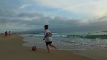 Ein Junge tritt bei Sonnenuntergang einen Fußball am Strand.