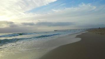 Kamerafahrt der Meereswellen, Strand und Pier bei Sonnenuntergang.