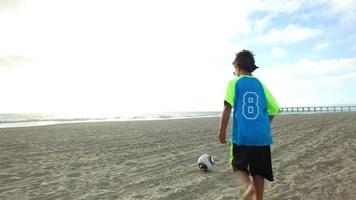 un ragazzo calcia un pallone da calcio sulla spiaggia al tramonto con l'oceano e il molo. video