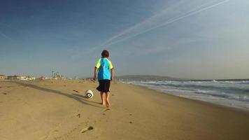 um menino chuta uma bola de futebol na praia ao pôr do sol.