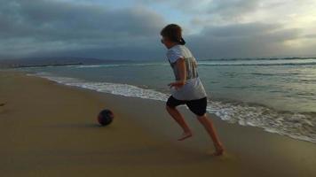 un ragazzo calcia un pallone da calcio sulla spiaggia al tramonto.