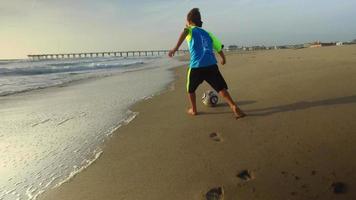 Ein Junge tritt bei Sonnenuntergang mit dem Meer und dem Pier einen Fußball am Strand.
