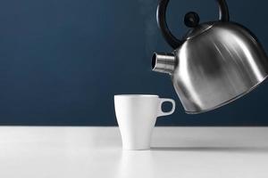 white mug with kettle on a white table. kettle pouring boiling water into a cup with smoke on wood table photo