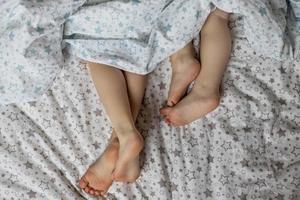 Close-up of two pairs of toddler girls feet on the bed under the blanket. Light blue and beige tones. photo