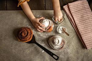 Children's hands in the frame sprinkles a fresh baked bun with cinnamon with pastry sprinkles photo