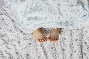 primer plano de los pies de la niña pequeña en la cama debajo de la manta. Tonos celestes y beige. foto