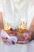 Manos de mujer sosteniendo un vaso de plástico transparente con mousse de chocolate, decorado con galletas en forma de conejito foto