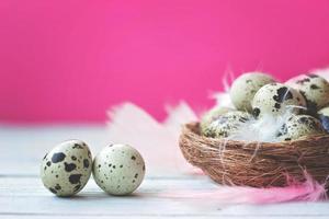 Huevos de codorniz en el nido con plumas de colores, sobre una mesa de madera blanca contra la pared rosa