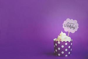 Cupcake de vainilla con pequeños corazones decorativos y cartel de feliz cumpleaños, en violeta taza de papel para hornear, contra el fondo violeta foto