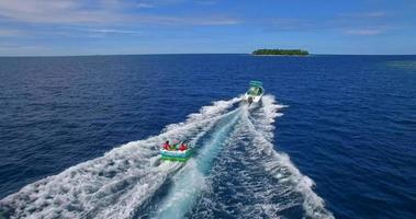 Vista aérea de un hombre y una mujer en un tubo inflable remolcando detrás de un bote a una isla tropical. video
