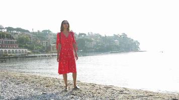 Una mujer camina en una playa con un vestido rojo mientras viaja en una ciudad turística de lujo en Italia, Europa.