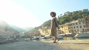 Una mujer caminando con un sombrero y una mochila viajando en Portofino, Italia, una ciudad turística de lujo en Europa. video